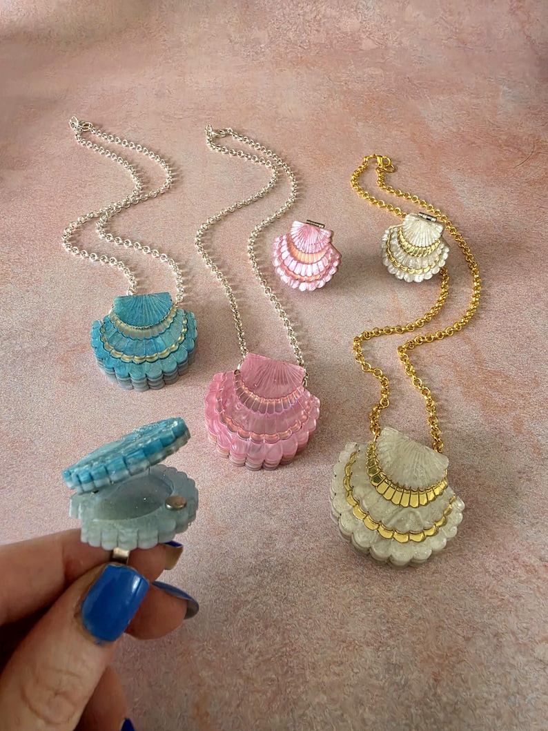 May include: A collection of shell-shaped lockets and rings in blue, pink, and white. The lockets hang on silver or gold chains, while the rings feature a hinged design that opens to reveal a small compartment. Each piece has a scalloped texture.