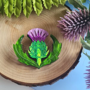 Pode incluir: Broche decorativo com um design estilizado de cardo. O broche tem uma flor roxa e rosa, folhas verdes e um botão verde e amarelo. O broche está sobre um fundo de madeira.