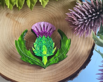 Puede incluir: Broche decorativo con un diseño de cardo estilizado. El broche tiene una flor de color púrpura y rosa, hojas verdes y un capullo verde y amarillo. El broche está sobre un fondo de madera.