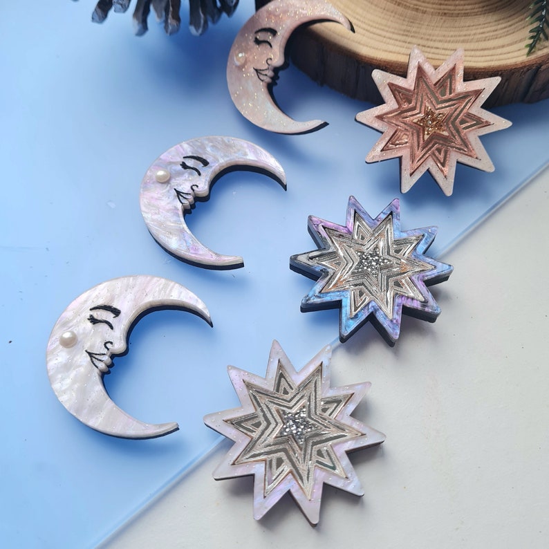 May include: Five decorative ornaments, two crescent moons and three stars, with iridescent finishes and pearl accents. The ornaments are made of wood and have a whimsical, celestial theme.