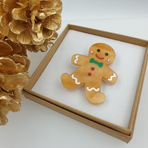 May include: A gingerbread man brooch with white icing details, red and green accents, and sparkly embellishments. The brooch is displayed in a brown box.