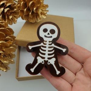 May include: A brown and white skeleton-shaped brooch. The brooch has a brown outline with a white skeleton design. The brooch is held in a hand, with a gold-painted pinecone and a brown box in the background.