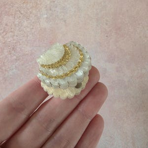 May include: A small, round, decorative trinket box shaped like a seashell. The exterior features a textured, off-white, pearlescent finish with horizontal ridges and a prominent, metallic gold band accenting the centre of the lid.