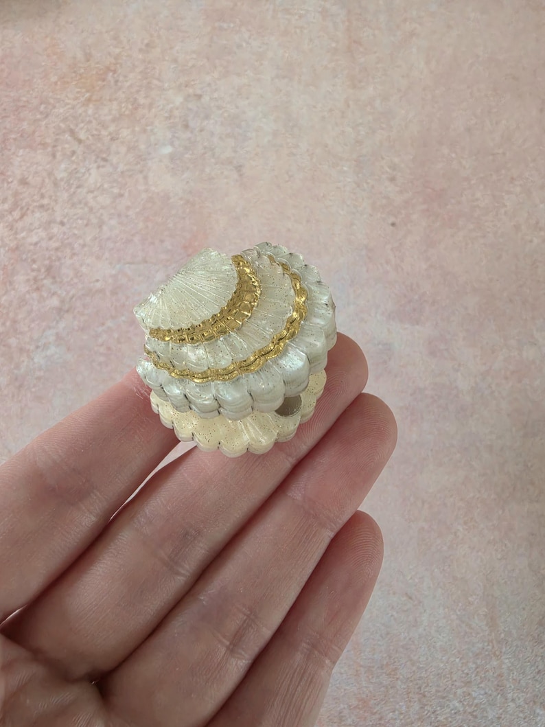 May include: A small, round, decorative trinket box shaped like a seashell. The exterior features a textured, off-white, pearlescent finish with horizontal ridges and a prominent, metallic gold band accenting the centre of the lid.