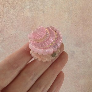 May include: A small, translucent pink seashell-shaped accessory with a ribbed texture and a scalloped edge. The base features a clear, glitter-infused layer with a small circular metallic component visible on the side, designed for attachment.