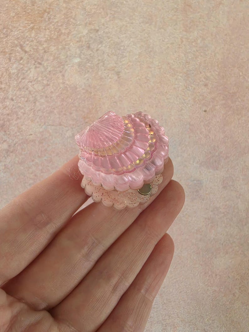 May include: A small, translucent pink seashell-shaped accessory with a ribbed texture and a scalloped edge. The base features a clear, glitter-infused layer with a small circular metallic component visible on the side, designed for attachment.