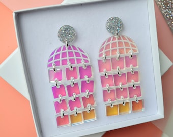 May include: A pair of iridescent pink and orange geometric earrings with silver accents. The earrings are in a white box with a white background.