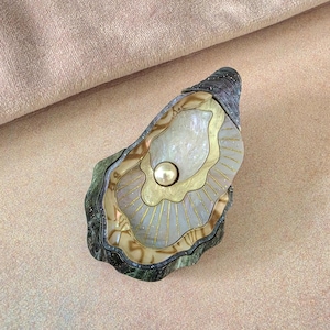 May include: A decorative oyster shell ornament featuring a dark, textured exterior and a polished interior. The center displays a smooth, cream-colored pearl set against a layered design of iridescent white and gold-toned accents with fine lines.