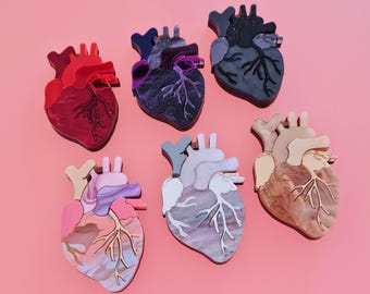 May include: Six anatomical heart shaped resin ornaments in various colors, including red, purple, black, white, and brown. The hearts are detailed with veins and chambers.