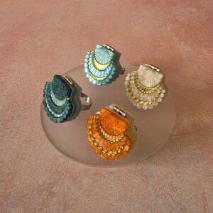 May include: Four rings featuring shell-shaped tops with hinged designs. Each ring displays a distinct colour: teal, light blue, cream, and orange. The shell surfaces are textured with fine lines and metallic gold accents along the scalloped edges.