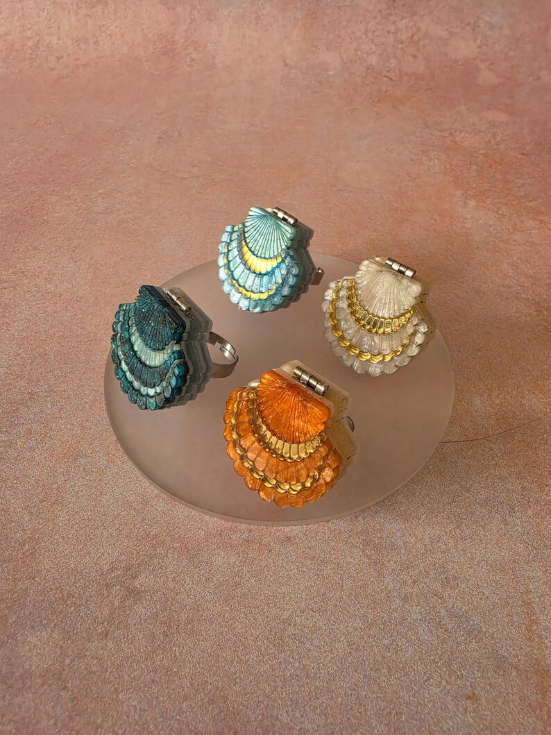 May include: Four rings featuring shell-shaped tops with hinged designs. Each ring displays a distinct colour: teal, light blue, cream, and orange. The shell surfaces are textured with fine lines and metallic gold accents along the scalloped edges.
