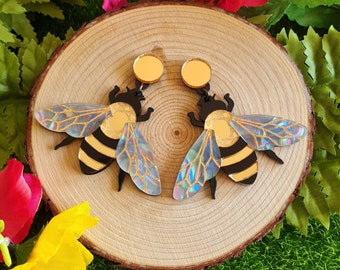May include: A pair of iridescent bee earrings with black and gold accents. The earrings are made of acrylic and have a gold-tone post.