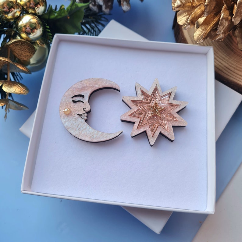May include: A white box with a pink and gold moon and star design. The moon has a smiling face and the star has a glittery center.