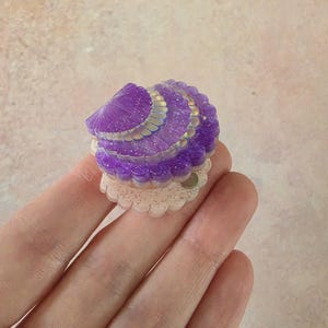 May include: A small, purple, translucent seashell-shaped accessory with a glittery finish. The top shell section features a scalloped edge with iridescent highlights, while the base is a pale, cream-coloured, scalloped circular piece.