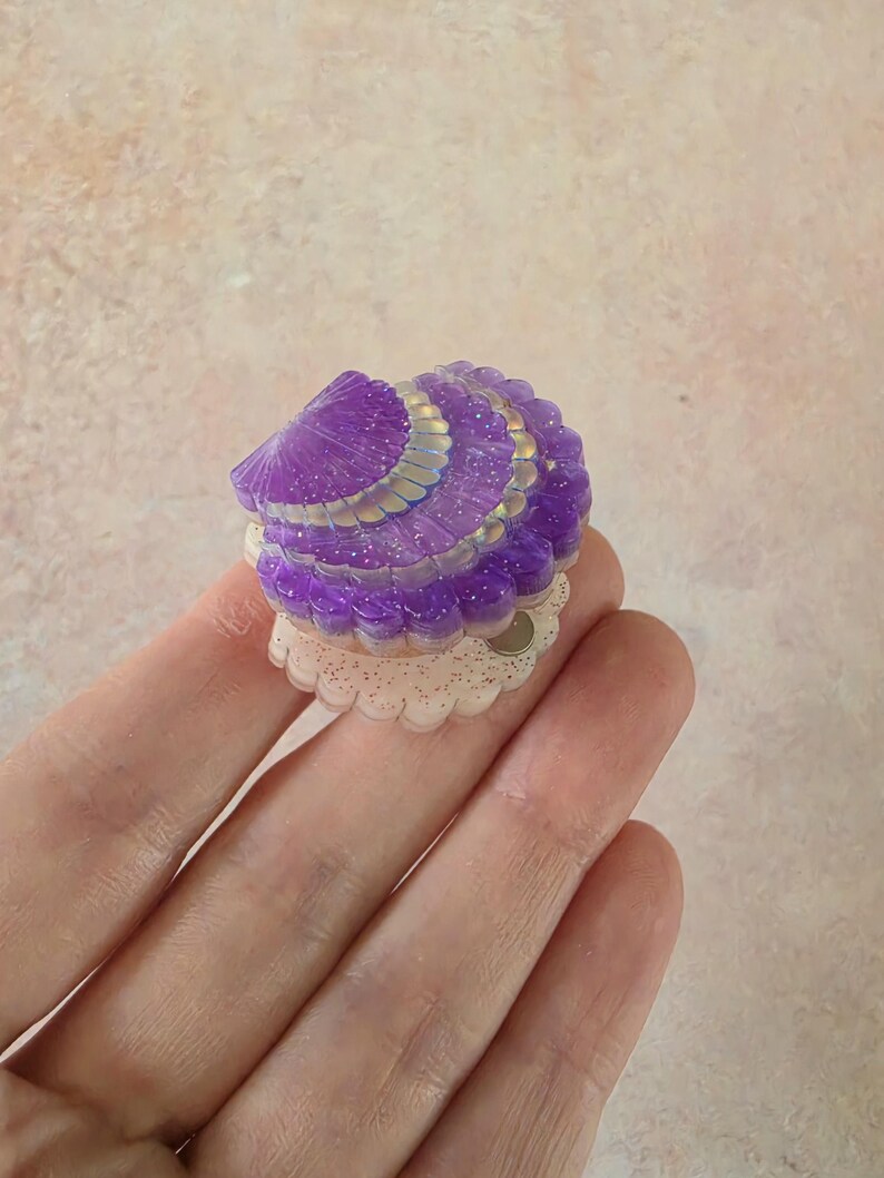 May include: A small, purple, translucent seashell-shaped accessory with a glittery finish. The top shell section features a scalloped edge with iridescent highlights, while the base is a pale, cream-coloured, scalloped circular piece.