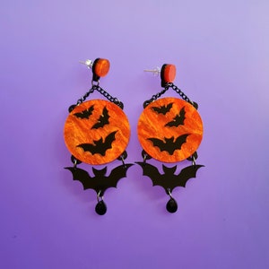 May include: A pair of black and orange Halloween-themed earrings with a dangling bat charm and a round orange disc with black bats.