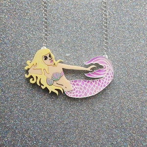 Puede incluir: Un collar con cadena plateada y un colgante de sirena rosa y morado. La sirena tiene el pelo rubio, un sujetador de concha verde y una cola rosa.