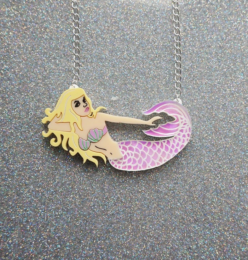 Puede incluir: Un collar con cadena plateada y un colgante de sirena rosa y morado. La sirena tiene el pelo rubio, un sujetador de concha verde y una cola rosa.