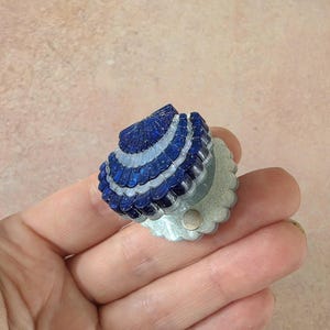 May include: A small, fan-shaped decorative object featuring alternating rows of deep blue and translucent white scalloped segments. The base is a pale, glittery off-white material with a small, circular metallic fastener visible on the front surface.