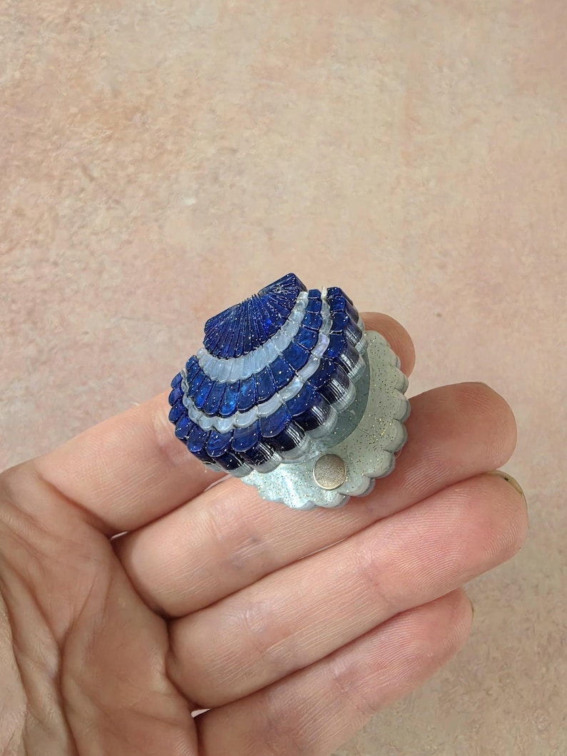 May include: A small, fan-shaped decorative object featuring alternating rows of deep blue and translucent white scalloped segments. The base is a pale, glittery off-white material with a small, circular metallic fastener visible on the front surface.