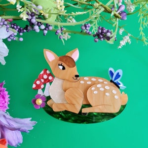 May include: A brown and white deer figurine with a blue butterfly on its back, lying on a green bed of leaves with red and white mushrooms behind it.