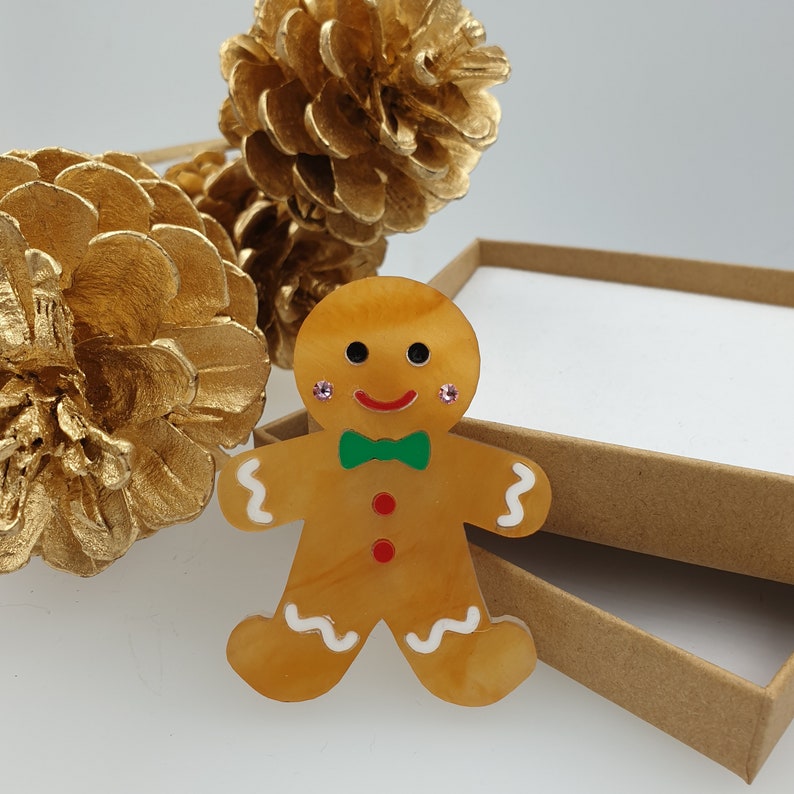 May include: A gingerbread man brooch with a green bow tie, red buttons, and pink glitter accents. The brooch is made of resin and has a glossy finish.