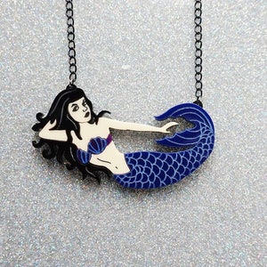 Puede incluir: Un collar de cadena negra con un colgante de sirena azul y blanco. La sirena est&aacute; tumbada de lado con la cola enroscada.