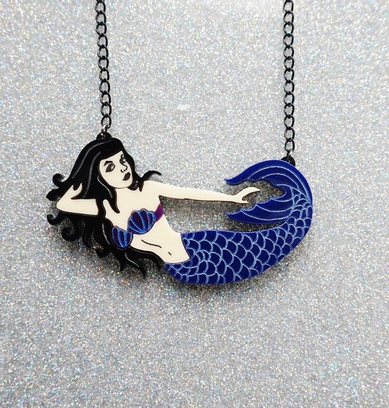 Puede incluir: Un collar de cadena negra con un colgante de sirena azul y blanco. La sirena est&aacute; tumbada de lado con la cola enroscada.