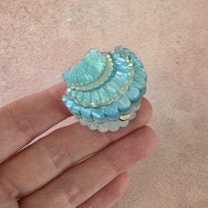 May include: A small, clam-shaped trinket box featuring a shimmering, iridescent blue and turquoise finish. The shell-like lid has layered, scalloped ridges with a glittery white base, creating a textured, multi-dimensional appearance.