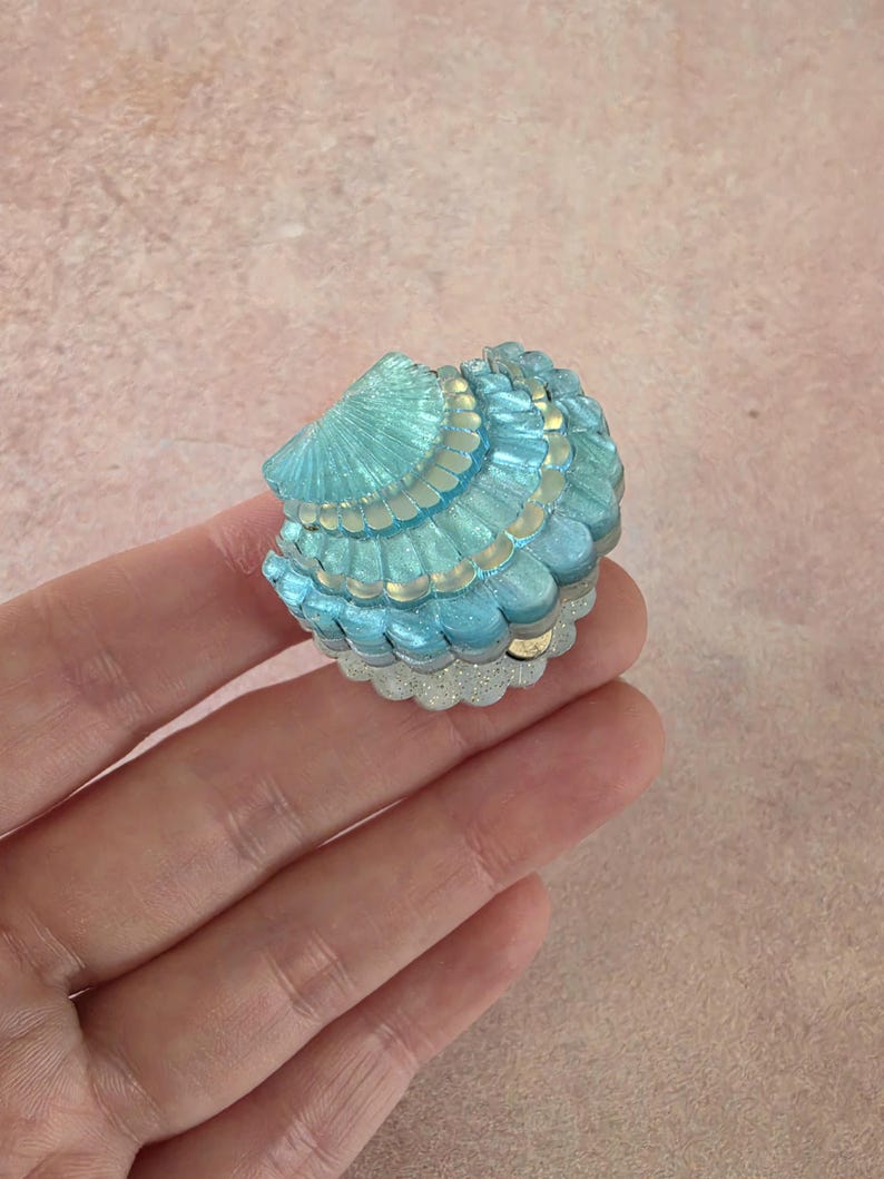 May include: A small, clam-shaped trinket box featuring a shimmering, iridescent blue and turquoise finish. The shell-like lid has layered, scalloped ridges with a glittery white base, creating a textured, multi-dimensional appearance.