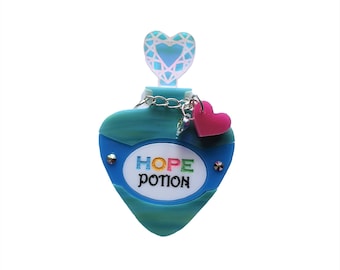Könnte beinhalten: Eine herzförmige Flasche in blaugrüner Farbe mit einer silbernen Kette und einem rosa Herz-Anhänger. Die Flasche hat ein weißes Etikett mit dem Text "HOPE POTION" in Regenbogenfarben.