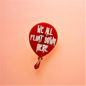 May include: Red balloon pin with a white inscription that reads "We all float down here". The balloon has a small red crystal hanging from the bottom.