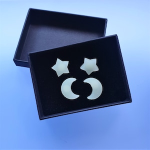 Puede incluir: Una caja de joyería de terciopelo negro con un par de pendientes de estrella y media luna que brillan en la oscuridad.