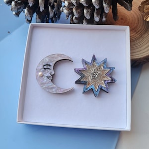 May include: A set of two wooden brooches in a white box. One brooch is a crescent moon with a pearl and a painted face. The other brooch is a multi-colored star with a glittery center.