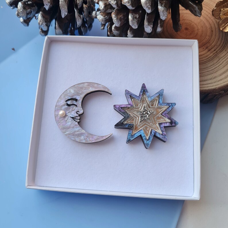 May include: A set of two wooden brooches in a white box. One brooch is a crescent moon with a pearl and a painted face. The other brooch is a multi-colored star with a glittery center.
