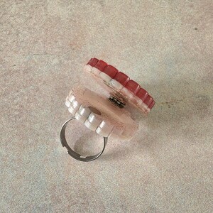 May include: A hinged ring featuring two circular, scalloped-edge components. The top piece is a deep red, while the bottom piece is a pale, glittery pink. The two sections are connected by a small metal hinge, and the base has a silver-toned band.