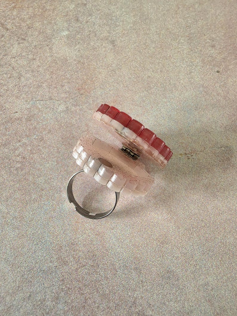 May include: A hinged ring featuring two circular, scalloped-edge components. The top piece is a deep red, while the bottom piece is a pale, glittery pink. The two sections are connected by a small metal hinge, and the base has a silver-toned band.