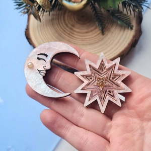 May include: A set of two pink and white wooden ornaments, a crescent moon with a face and a star with a glitter center.