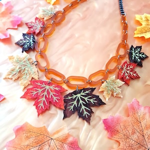 May include: A brown and orange necklace with a chain link design and multiple fall-themed maple leaf charms. The leaves are in shades of red, orange, green, and brown.