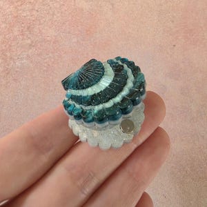 May include: A small, shell-shaped accessory featuring a gradient of deep teal to light blue with gold glitter accents. The top portion is a dark, ridged shell, while the bottom base is a translucent, glittery white scalloped edge with a small metal pin.