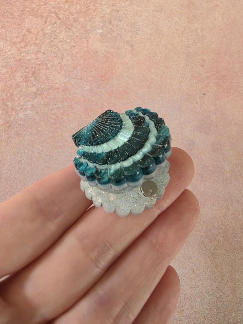 May include: A small, shell-shaped accessory featuring a gradient of deep teal to light blue with gold glitter accents. The top portion is a dark, ridged shell, while the bottom base is a translucent, glittery white scalloped edge with a small metal pin.