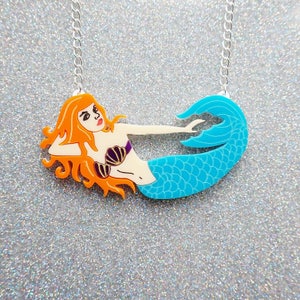Puede incluir: Un collar de sirena turquesa y naranja con una cadena plateada. La sirena est&aacute; tumbada de lado con la cola enroscada. Tiene el pelo largo y rojo y lleva un sujetador de concha de mar morado.