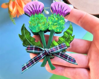 Puede incluir: Un broche de cardo verde y morado con un lazo de cinta a cuadros. El broche está hecho de resina y tiene un acabado brillante.