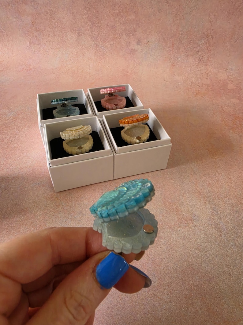 May include: A collection of small, clam-shaped resin trinket boxes with hinged lids. Each box features a unique colour, including blue, pink, white, and orange. The boxes are presented in individual white square gift boxes with black inserts.