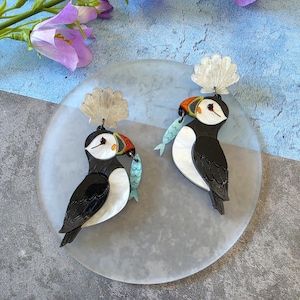 May include: A pair of dangle earrings featuring puffin birds with black and white bodies, orange and black beaks, and a small light blue fish held in each beak. Each puffin is topped with a translucent, scalloped shell-shaped accent piece.
