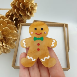 May include: A gingerbread man brooch made of acrylic with a green bow tie and pink rhinestones for eyes. The gingerbread man is a light brown color with white icing details.