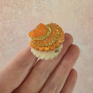 May include: A small, round, clam-shaped trinket box featuring a shimmering orange lid with gold-toned scalloped detailing and a textured, off-white base. The lid has a fan-like pattern with ridges, and the base has a repeating vertical ribbed design.