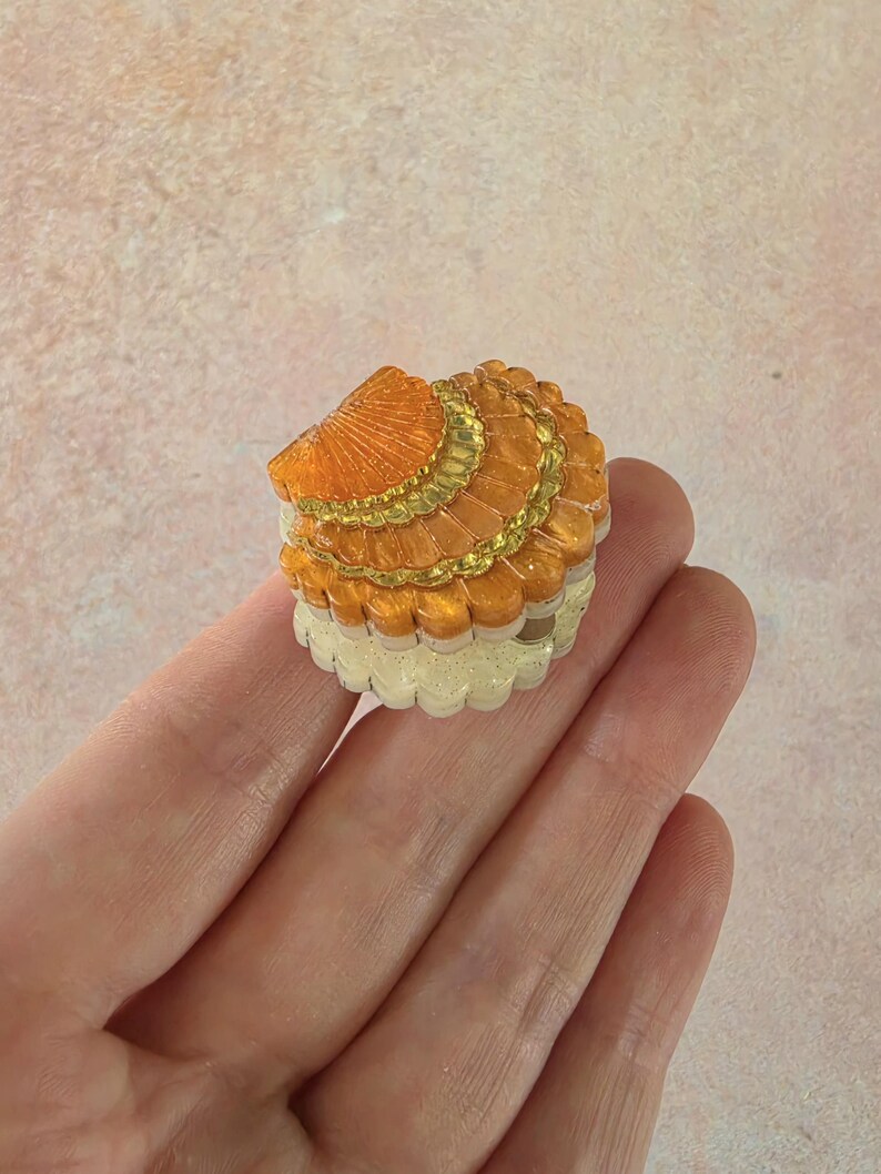 May include: A small, round, clam-shaped trinket box featuring a shimmering orange lid with gold-toned scalloped detailing and a textured, off-white base. The lid has a fan-like pattern with ridges, and the base has a repeating vertical ribbed design.