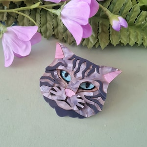 Puede incluir: Un broche de gato gris y blanco con ojos azules y nariz rosa. El broche está hecho de resina y tiene un diseño detallado.