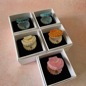May include: Five small, open shell-shaped trinket boxes in individual white square boxes with black inserts. The shells come in blue, light blue, white with gold trim, orange, and pink, each featuring a textured surface and a small magnetic closure.